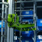 Zielone ramię robota sortuje niebieskie skrzynki w zautomatyzowanym magazynie